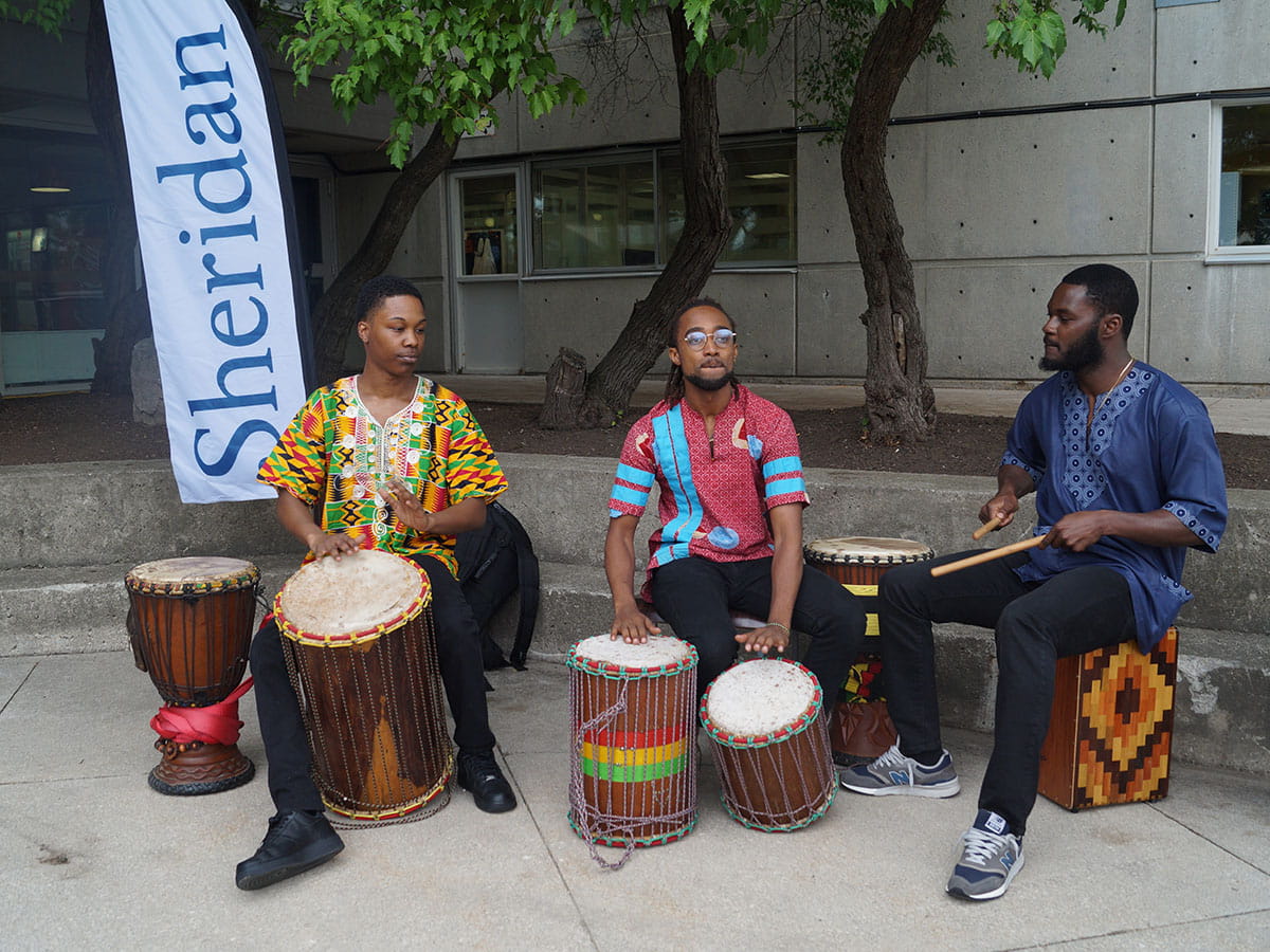 Three people playing drums outside. A Sheridan flag can be seen in the background.