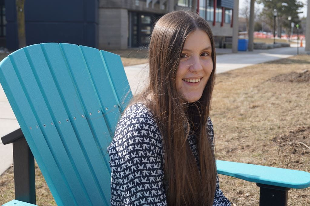 Mikayla Bianchin smiling outside sitting on a blue chair on Sheridan's campus