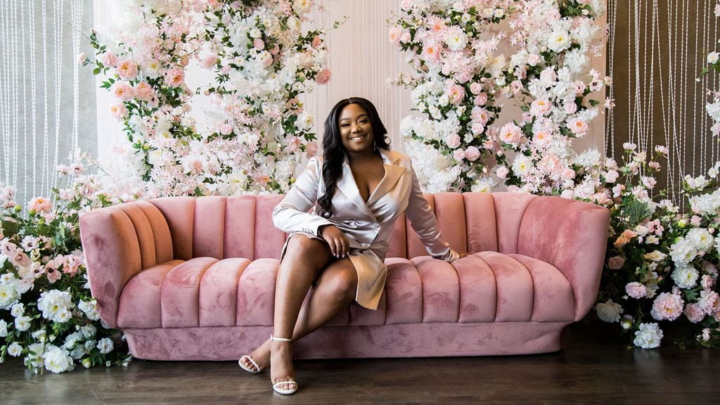 Pauleanna Reid smiling while sitting on a pink sofa surrounded by flowers.