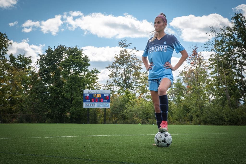 Woman standing in a soccer uniform on a field with hands on hips and left foot on a soccer ball
