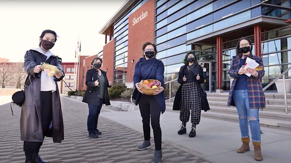 The Putting Food on the Table Research Team wearing masks and holding food while standing in front of a building at Sheridan's Trafalgar Road Campus.