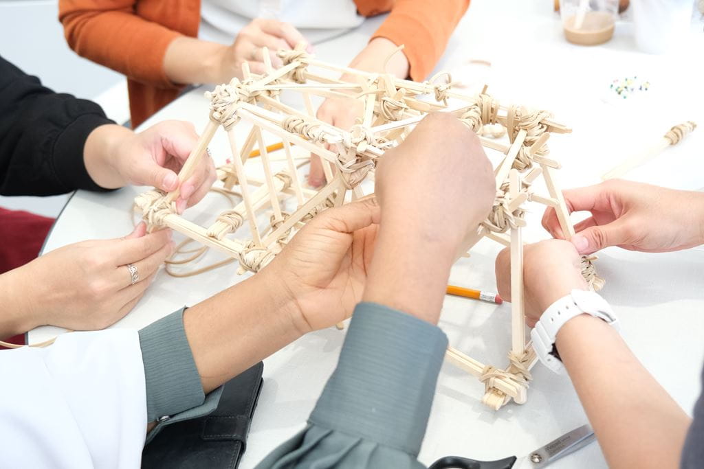 Hands of several different Sheridan architecture students are pictured building a structure out of popsicle sticks