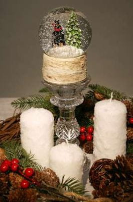 A holiday tabletop display comprised of a snow globe, candles and greenery with pinecones and berries