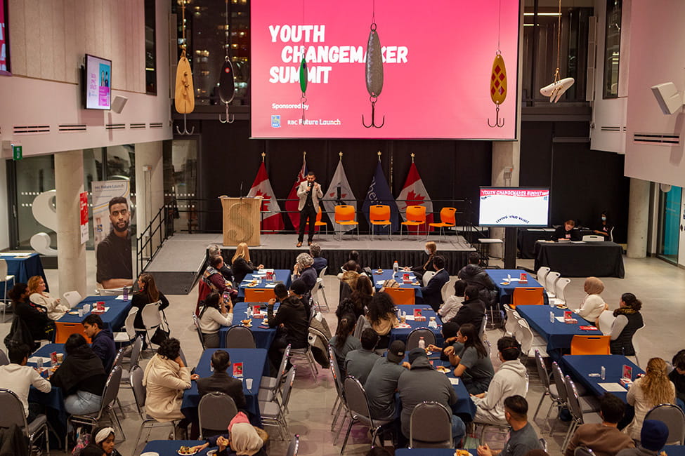 People seated around tables looking at a stage. A sign about the stage reads 'Youth Changemaker Summit Sponsored by RBC Future Launch'