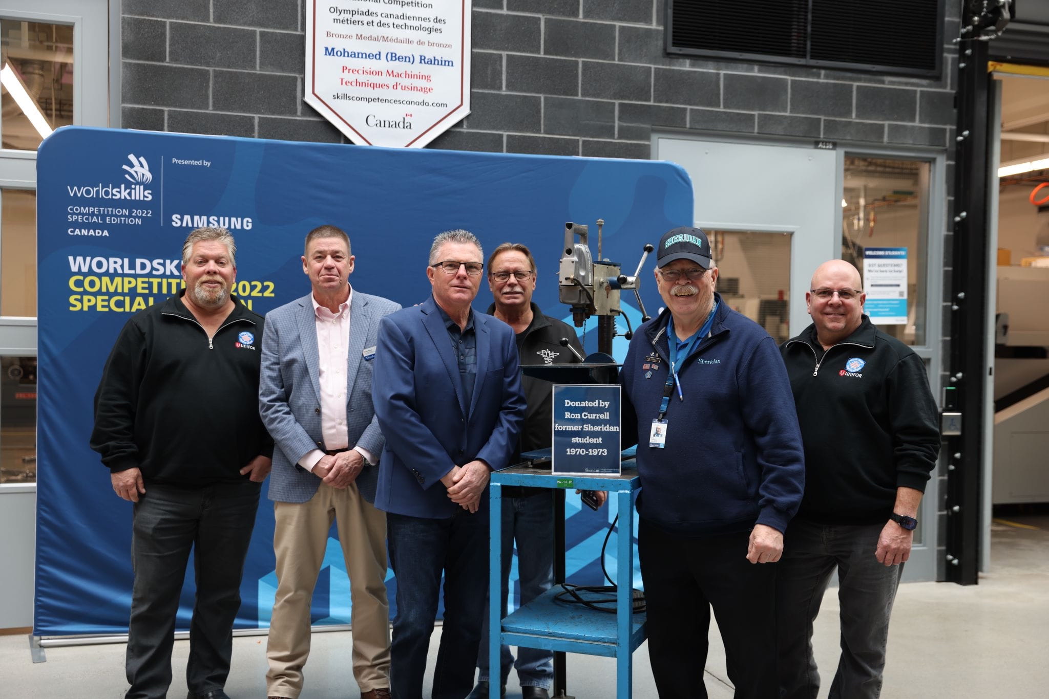 Sheridan graduate Ron Currell stands beside a drill press he donated to the college, surrounded by members of Unifor and Magna School for the Skilled Trades Associate Dean Greg Royal.