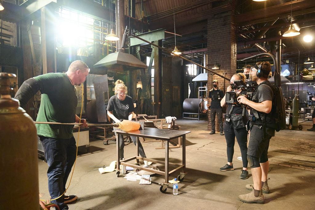Glassworkers shape a piece of glass while a film crew captures the action on the set of Blown Away